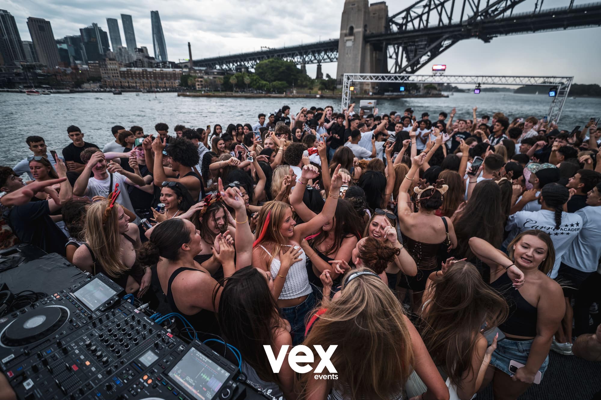 A crowd dancing around DJ decks on a boat beneath the Sydney Harbour Bridge in daylight