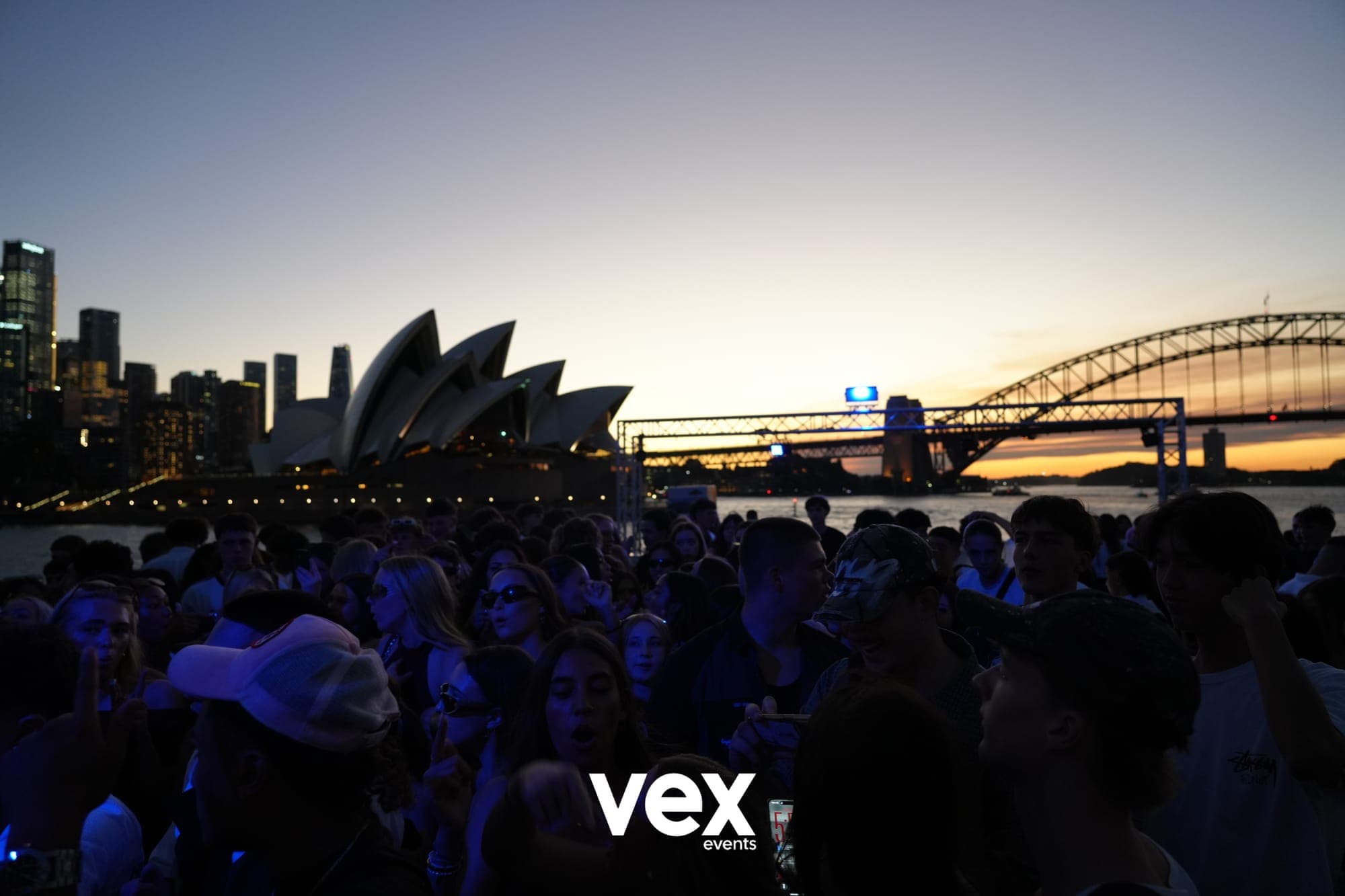 A blue-lit boat crowd at dusk with the Sydney Opera House and Harbour Bridge behind them