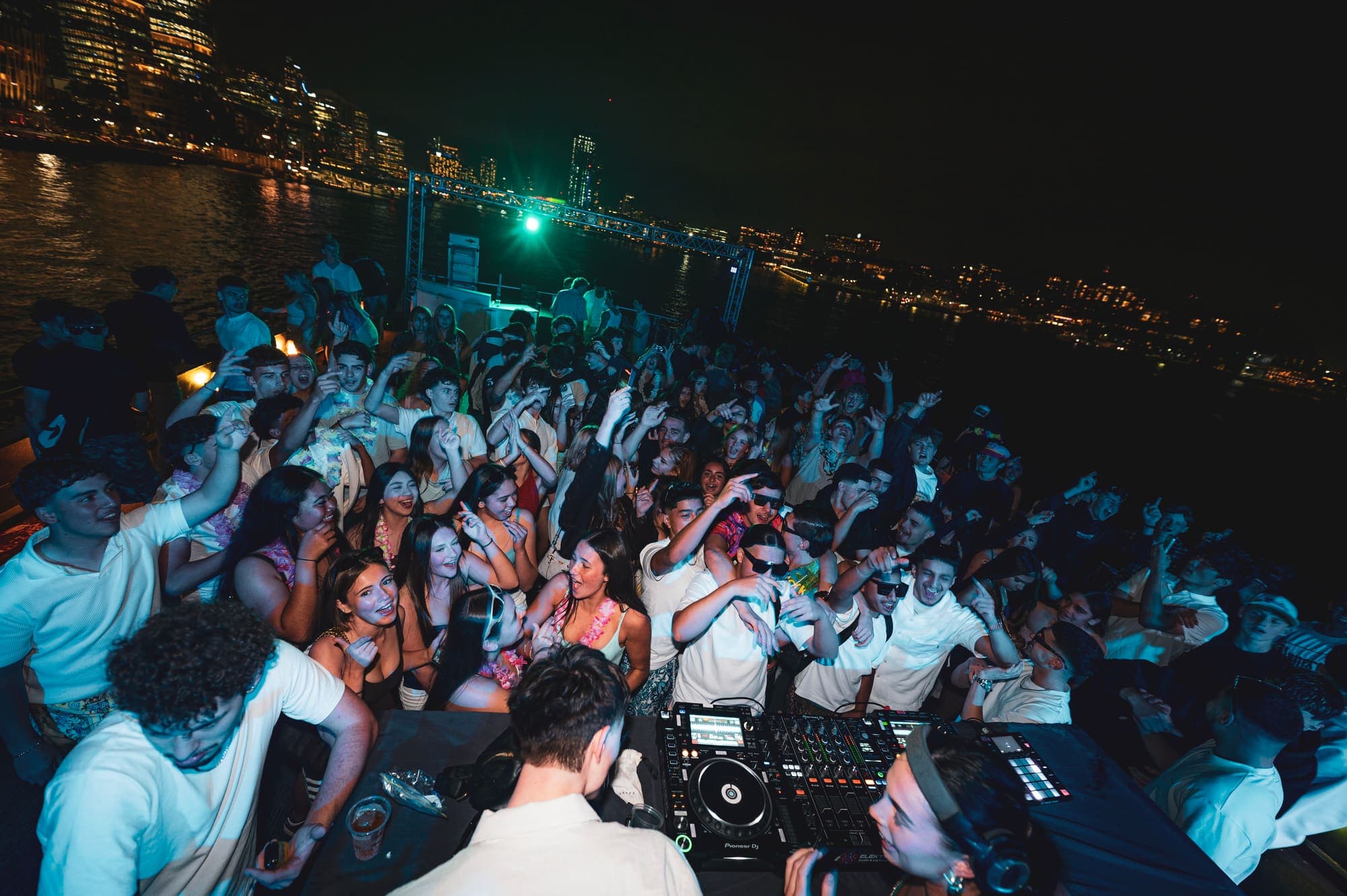 A wide view over DJ decks towards a blue-lit crowd, with city lights across the water at night